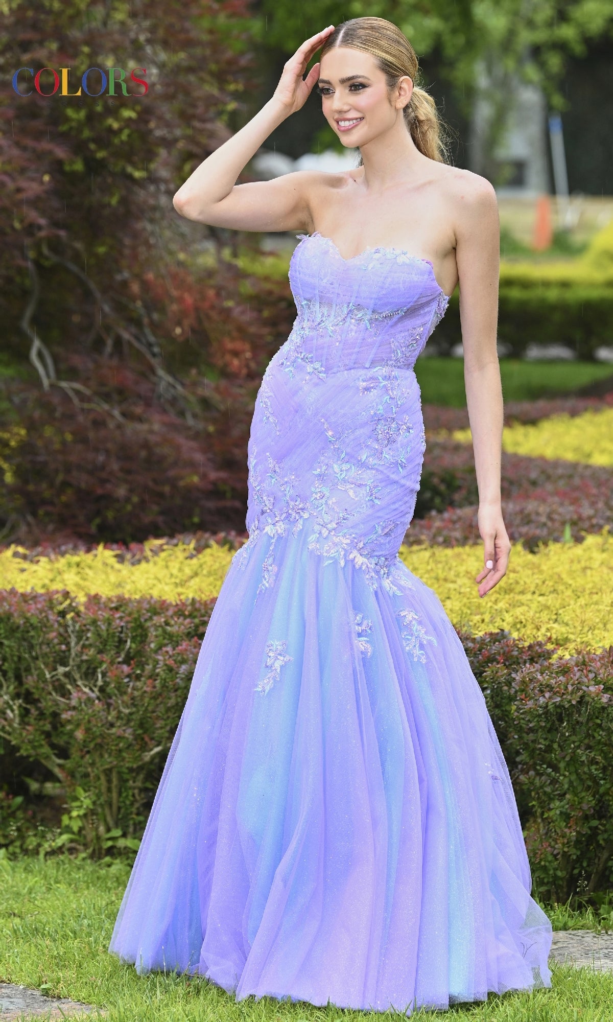 A woman smiles and touches her hair while standing on grass, wearing the Colors Dress 3529—an embroidered, strapless mermaid prom dress.